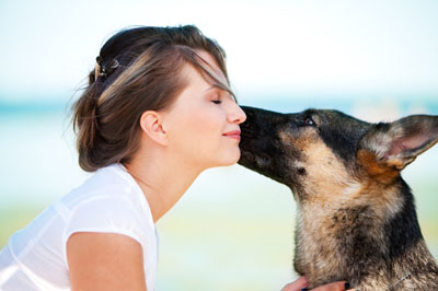 A woman with her eyes closed and a smile on her face as a German Shepherd licks her cheek, photographed outdoors against a pale sky.