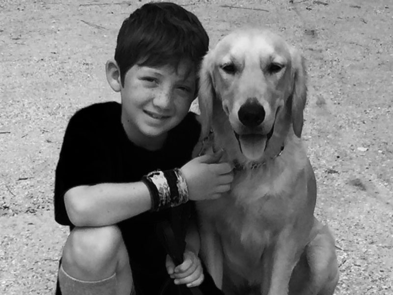 A black and white photograph of a boy crouching outdoors with his arm around a light coated dog.