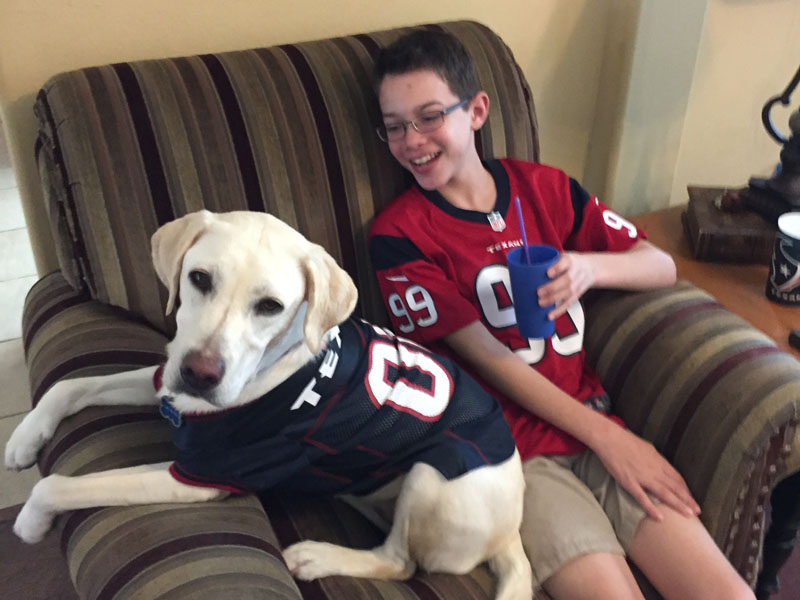 A boy in a football jersey sitting on a sofa holding a drink, with a pale yellow Labrador in a matching jersey lying against him.
