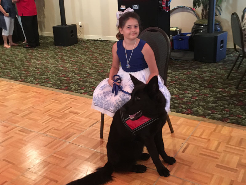 A girl in a blue and white dress sits on a chair at the edge of a parquet dance floor with a black German Shepherd in a red harness sitting at her feet.