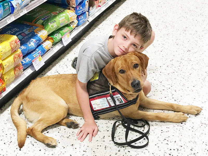 A boy kneels in a supermarket pet food aisle with both arms around a tan dog lying beside him in a black service dog vest with red ID panels.
