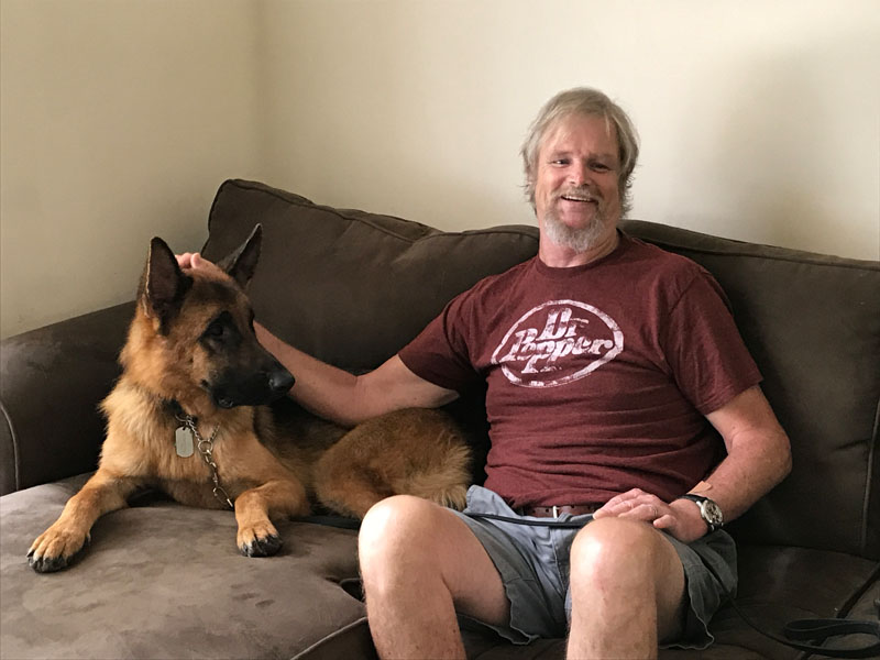 A man sits on a brown couch at home with his hand resting on the head of a German Shepherd lying beside him, a leash across his lap.