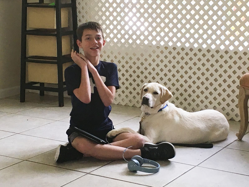 A boy sitting cross legged on a tile floor with a yellow Labrador lying beside him and a pair of headphones on the floor.