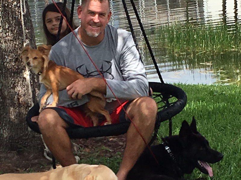 A man sits in a round garden swing beside a pond with a small tan dog on his lap, a girl standing behind the swing, and a black German Shepherd and a yellow dog lying on the grass in front.
