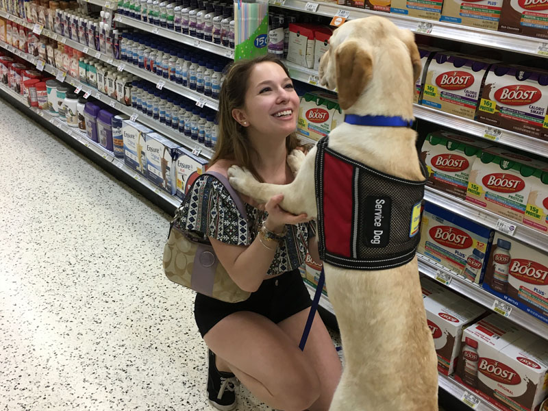 A woman crouching in a supermarket aisle laughing as a yellow Labrador in a red Service Dog vest puts its front paws in her hands.