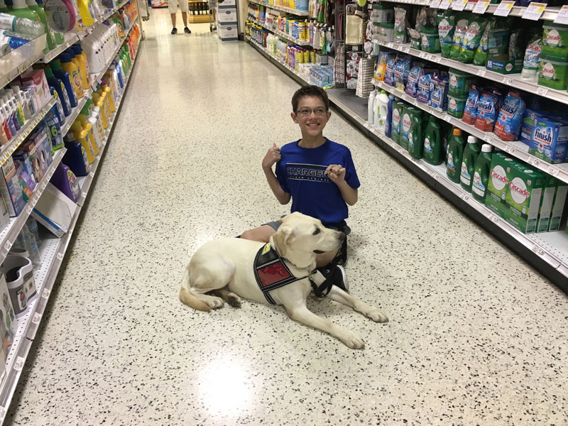 A boy kneeling in a supermarket cleaning aisle giving two thumbs up beside a yellow Labrador lying calmly in a service dog vest.