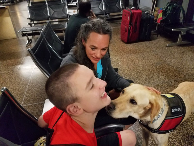 In an airport waiting area, a young man seated in a wheelchair tips his head back toward a yellow Labrador in a red vest patched Service Dog, while a woman crouches behind them beside the luggage.